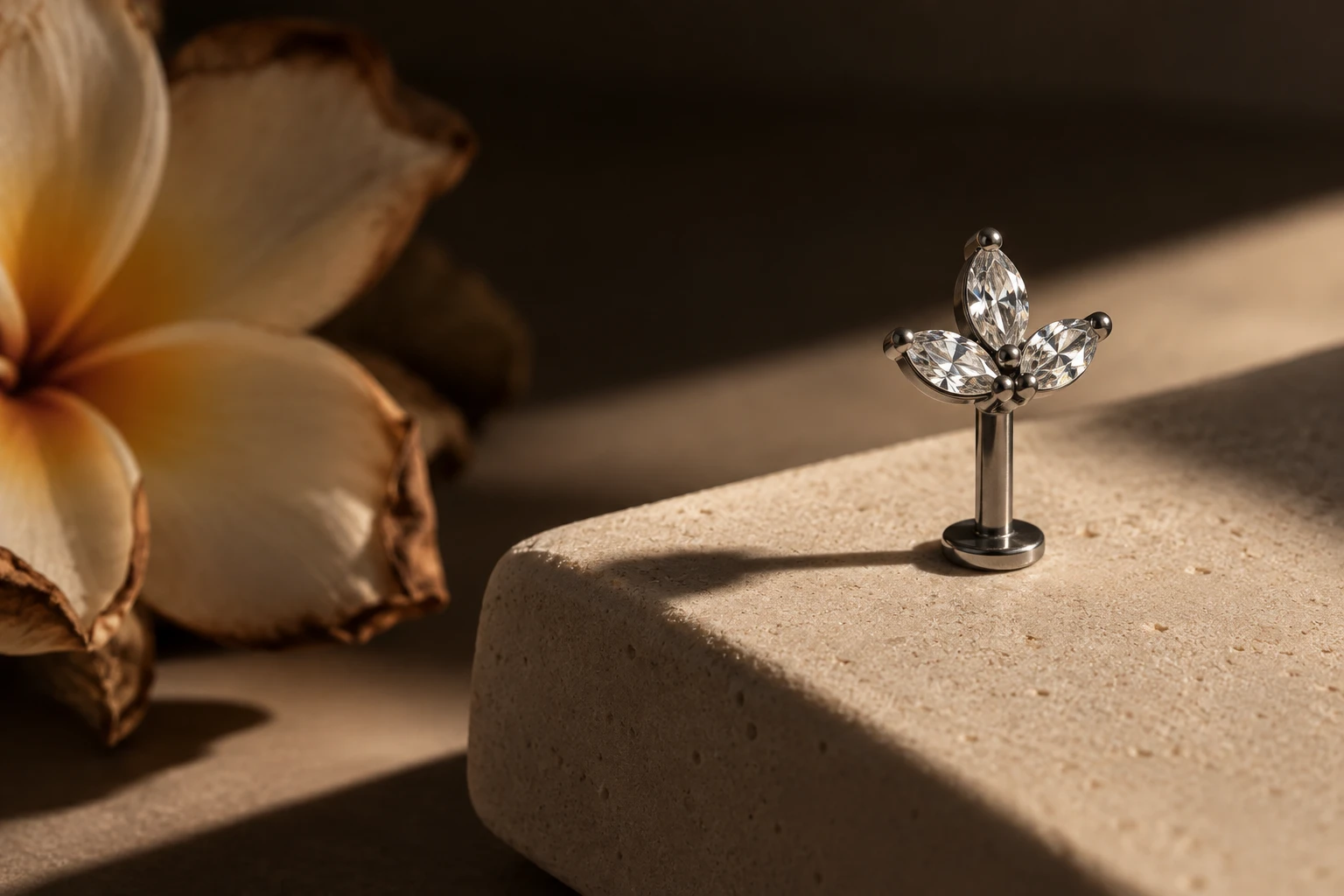 Top fleur en titane à trois cristaux taille marquise, sur une pierre claire, fleur de frangipanier en arrière-plan